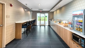 A modern kitchen area with a long counter, bar stools, a refrigerator stocked with beverages, and a view of the outdoors through the door. - Microtel Inn & Suites by Wyndham Farmington (Farmington)