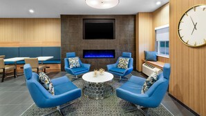 A modern lounge area with blue chairs around a central table, a flat-screen TV mounted on the wall, and a built-in fireplace. - Microtel Inn & Suites by Wyndham Farmington (Farmington)