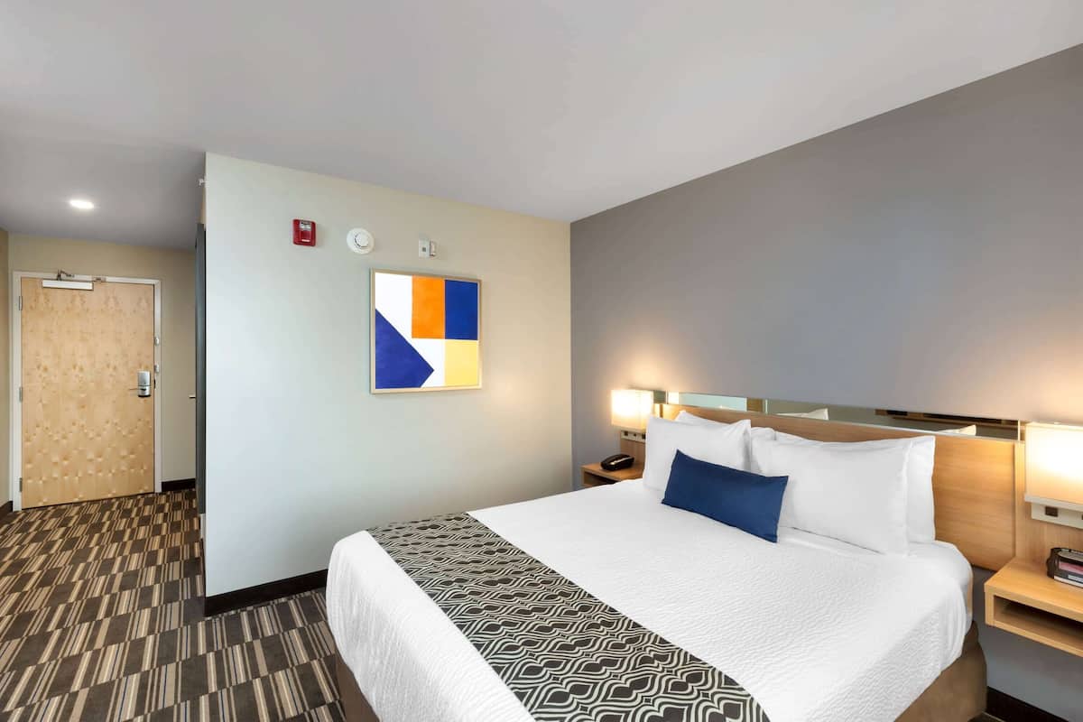 A hotel room with a large bed, a bedside table with a lamp, a wall clock, and a fire alarm.