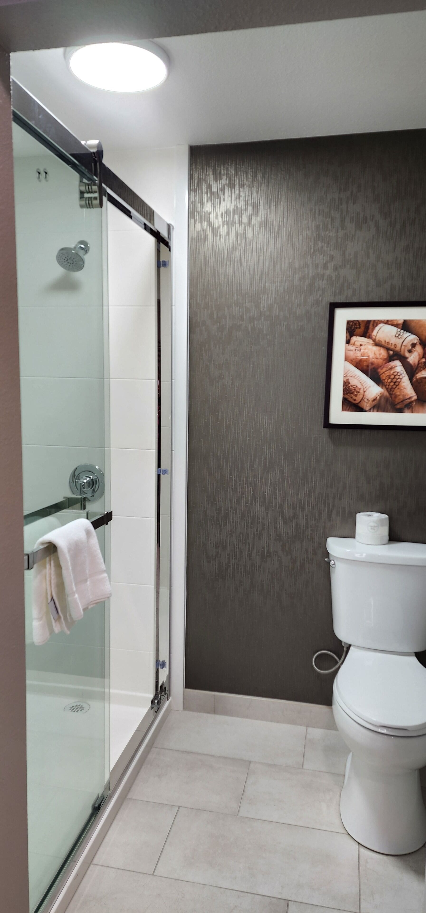 Une salle de bain moderne avec une douche à parois de verre, des toilettes blanches et un tableau encadré représentant des bouchons de bouteilles de vin, accroché au mur.