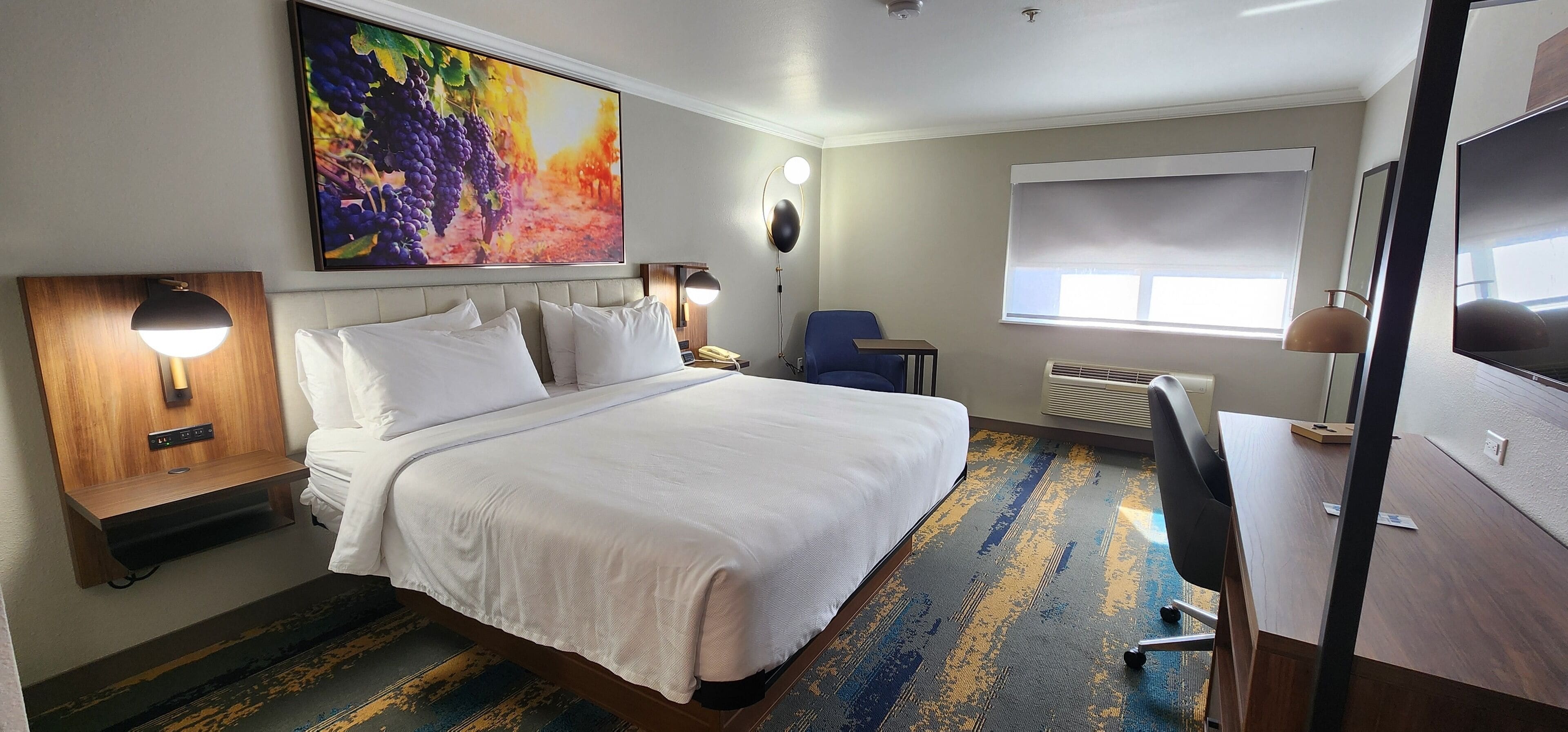 Ein Hotelzimmer mit einem großen Bett, einem Schreibtisch, einem Stuhl, einem Fernseher und einem Gemälde an der Wand.