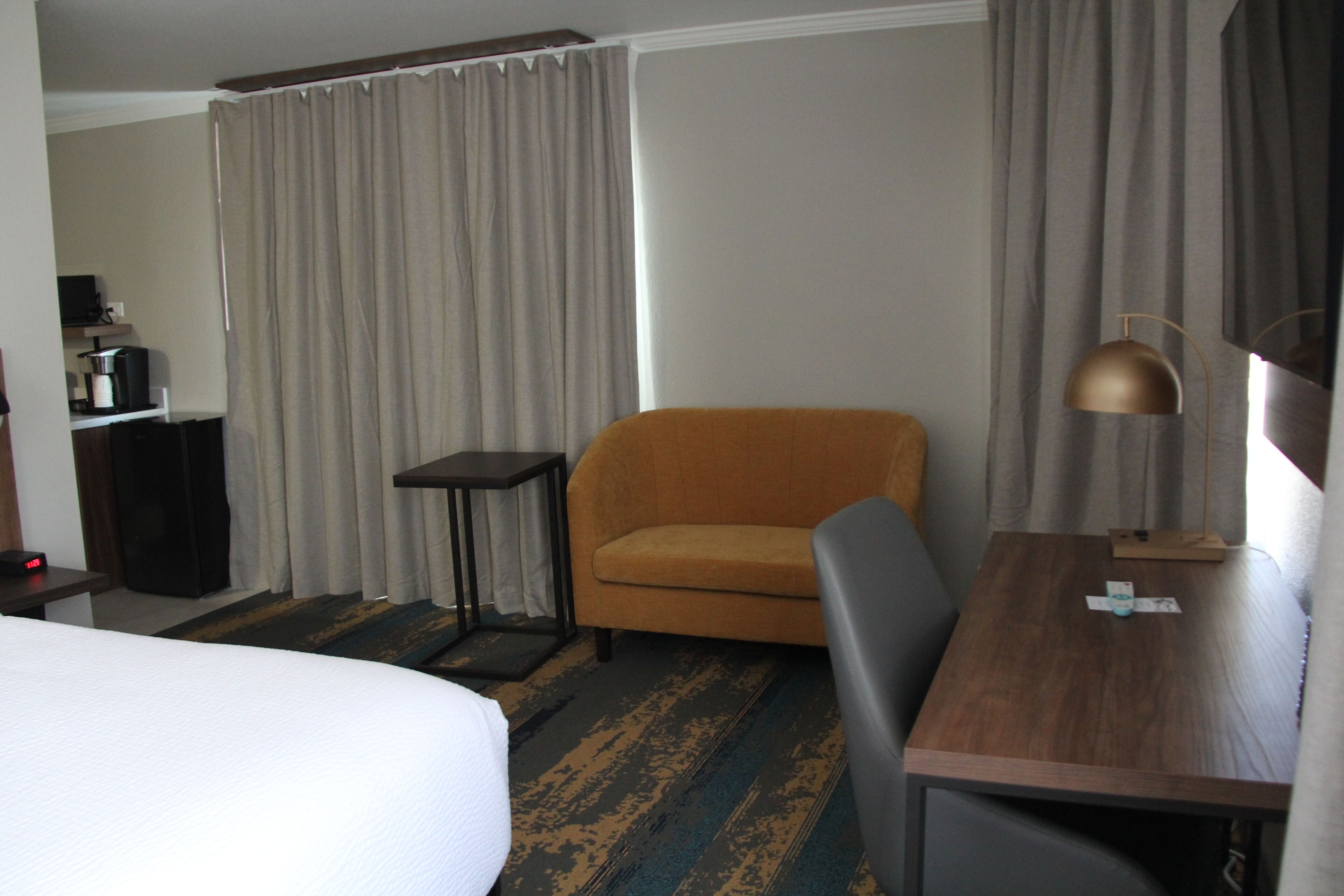 Une chambre d’hôtel avec un lit, une petite table avec une lampe, une chaise, un bureau et un rideau.