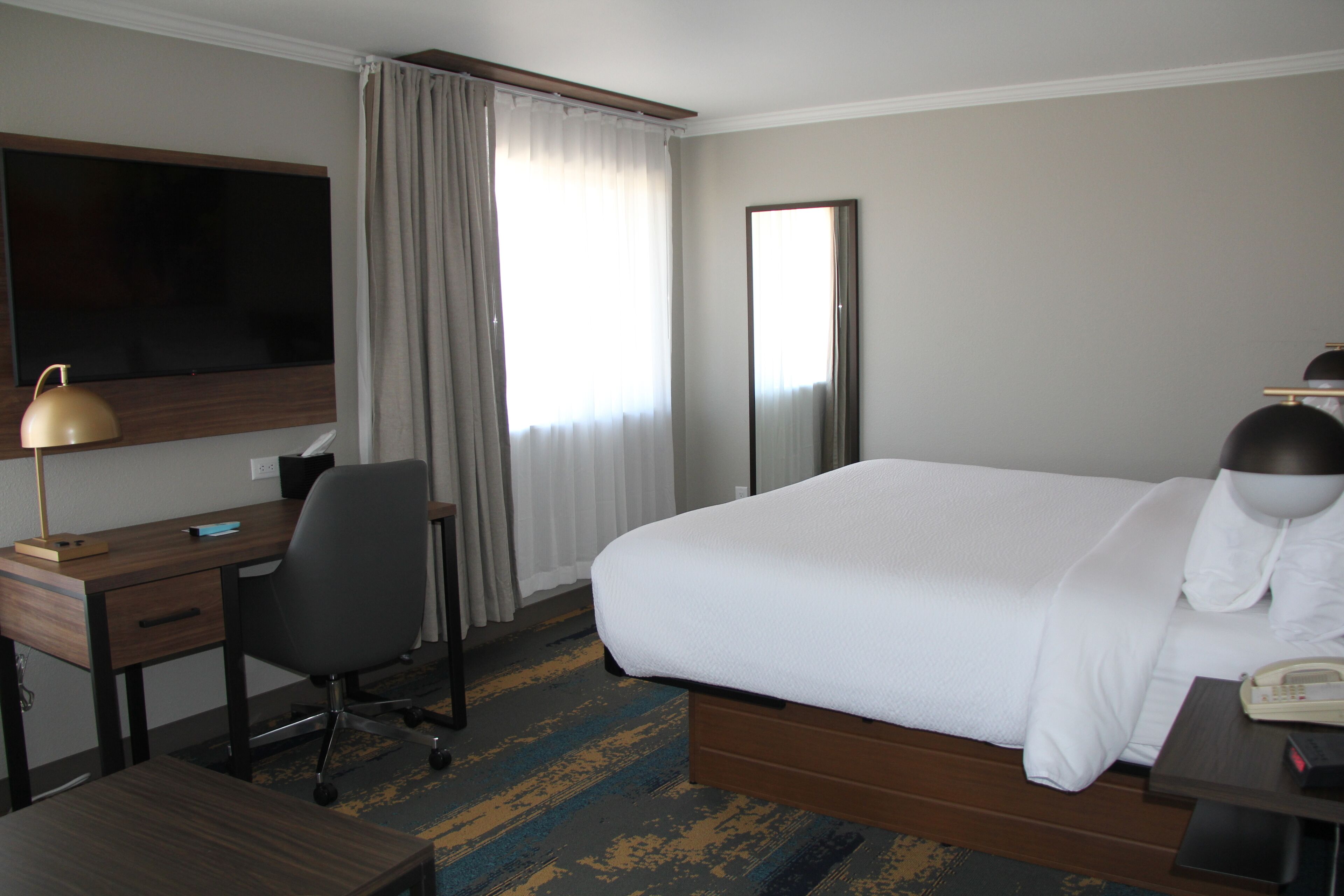 Une chambre d’hôtel équipée d’un lit, d’un bureau, d’une chaise, d’une télévision, d’un téléphone et d’une fenêtre avec des rideaux.