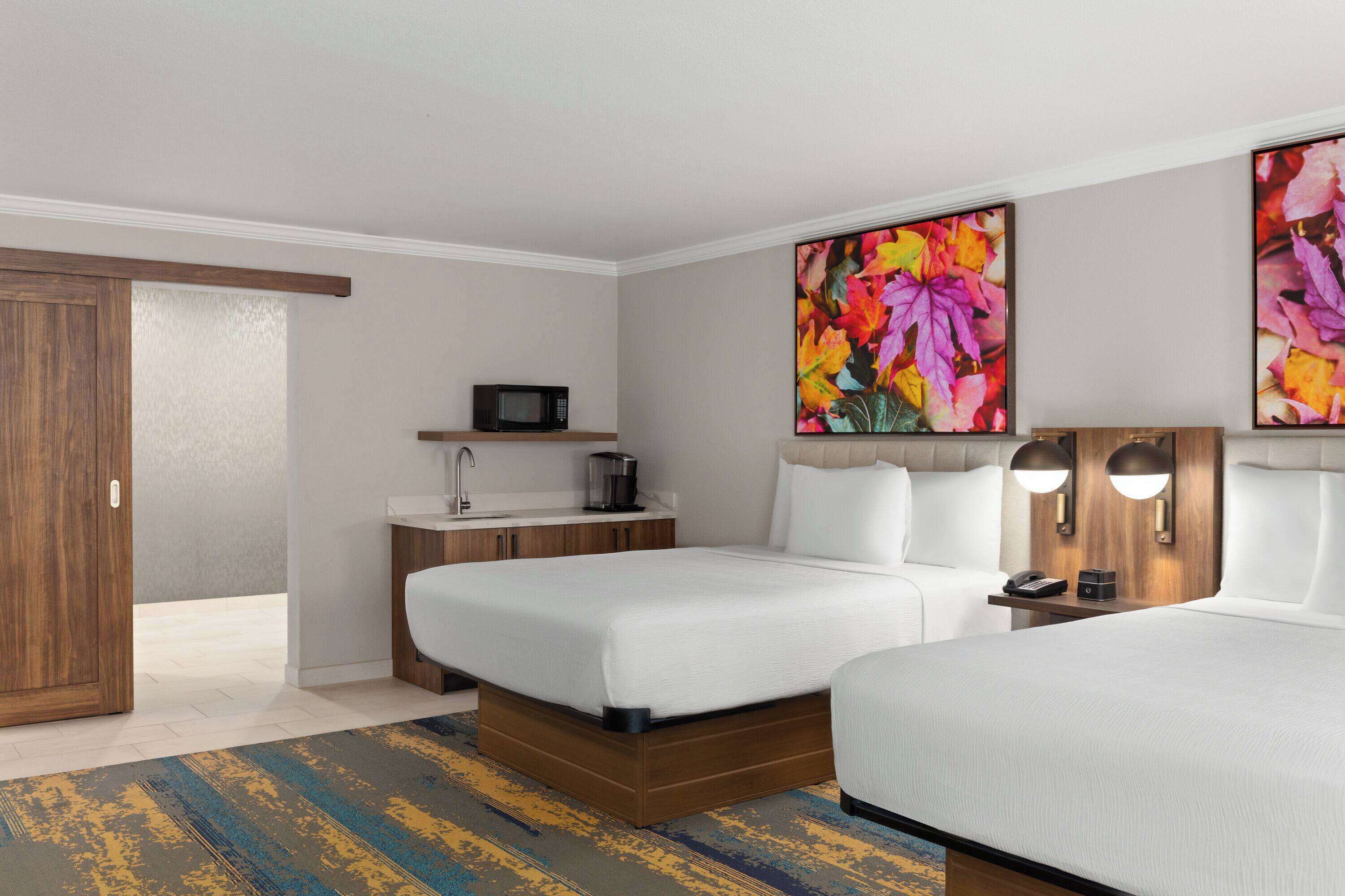 A hotel room with two beds, a microwave, a small table with a lamp, and artwork on the wall.