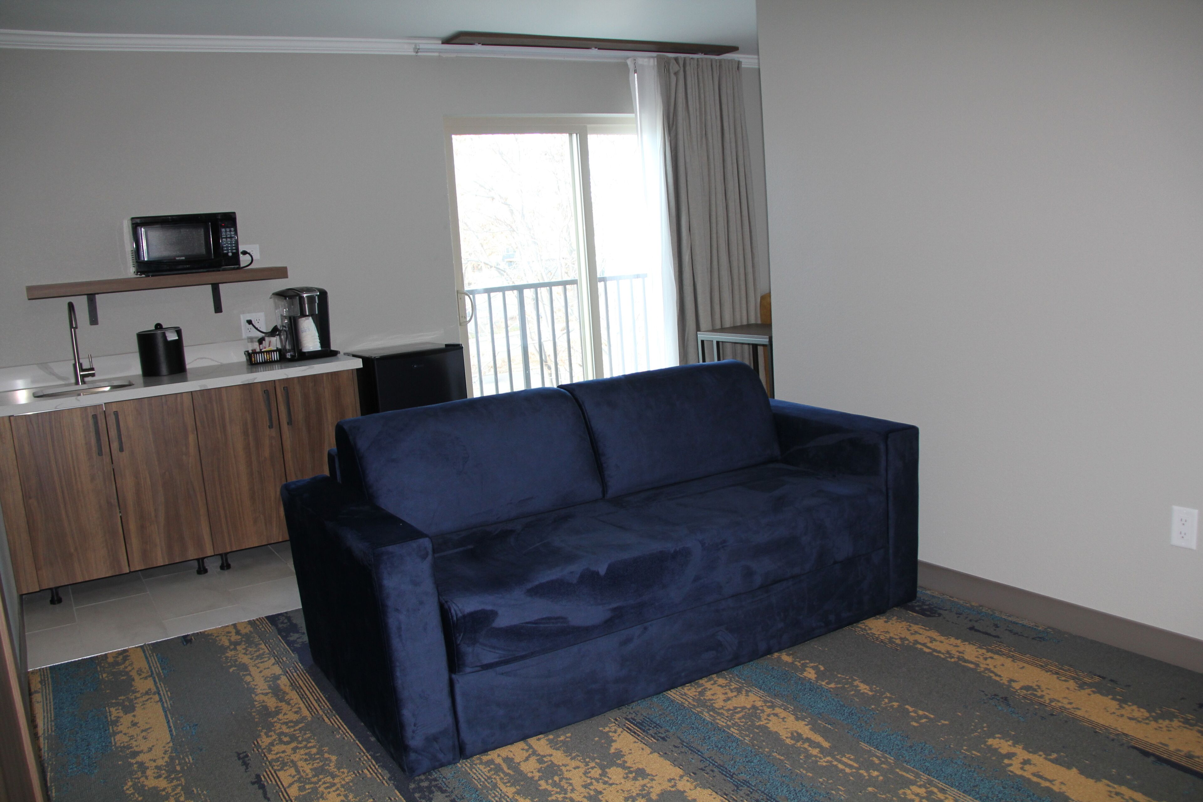 A compact living space with a dark blue sofa, a kitchenette with a microwave, coffee maker, and a small table, and a sliding glass door leading to a balcony.