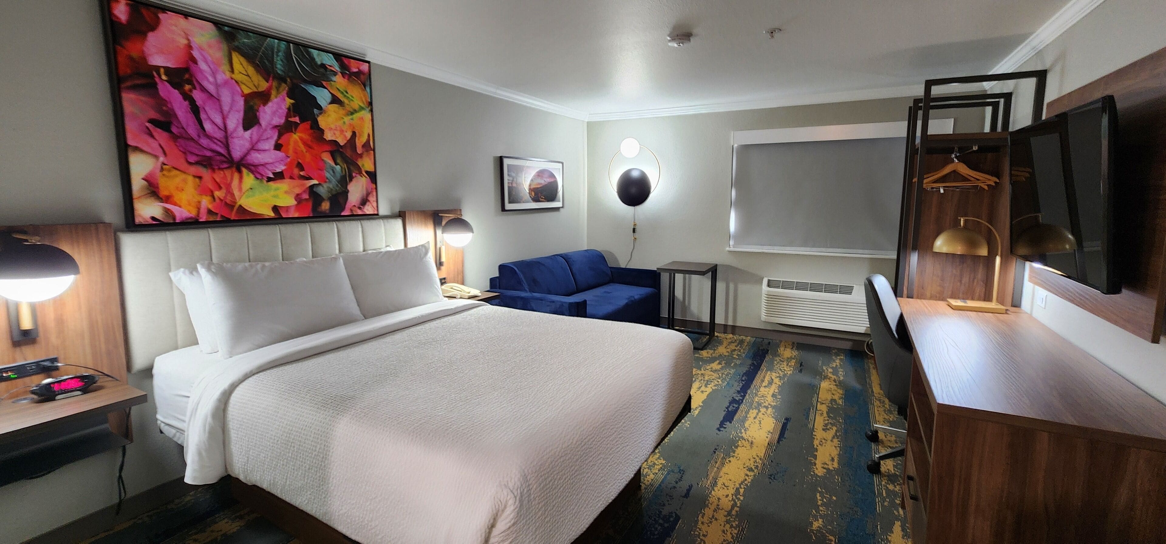 A hotel room with a large bed, a desk with a lamp, a blue chair, a TV, and a painting on the wall.