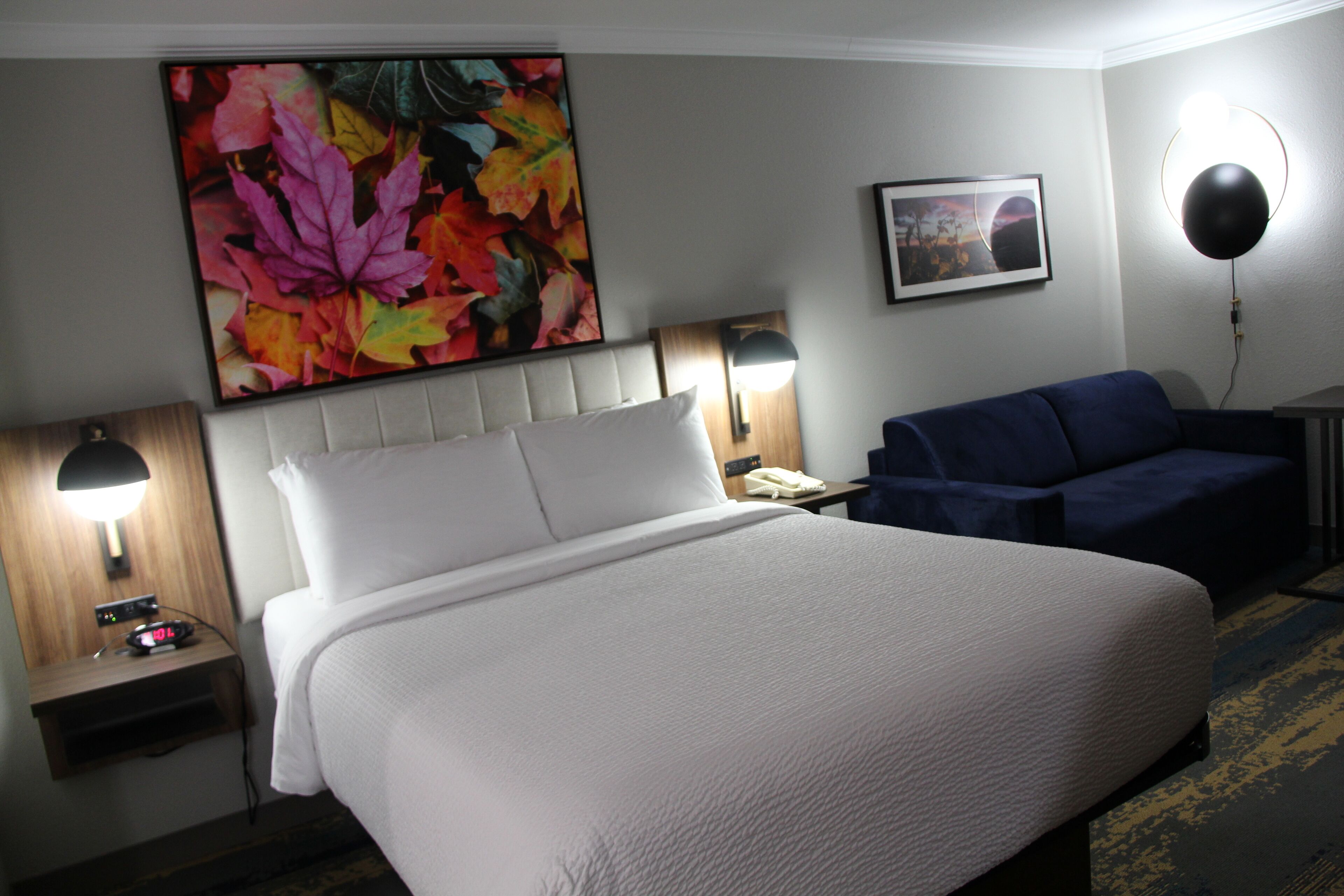 A hotel room with a large bed, two bedside tables, a desk, a chair, a wall-mounted lamp, and a framed picture.