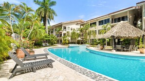 A resort pool area with lounge chairs, a thatched-roof structure, and a multi-story building with balconies. - Beachfront Jewel in Langosta (tamarindo)