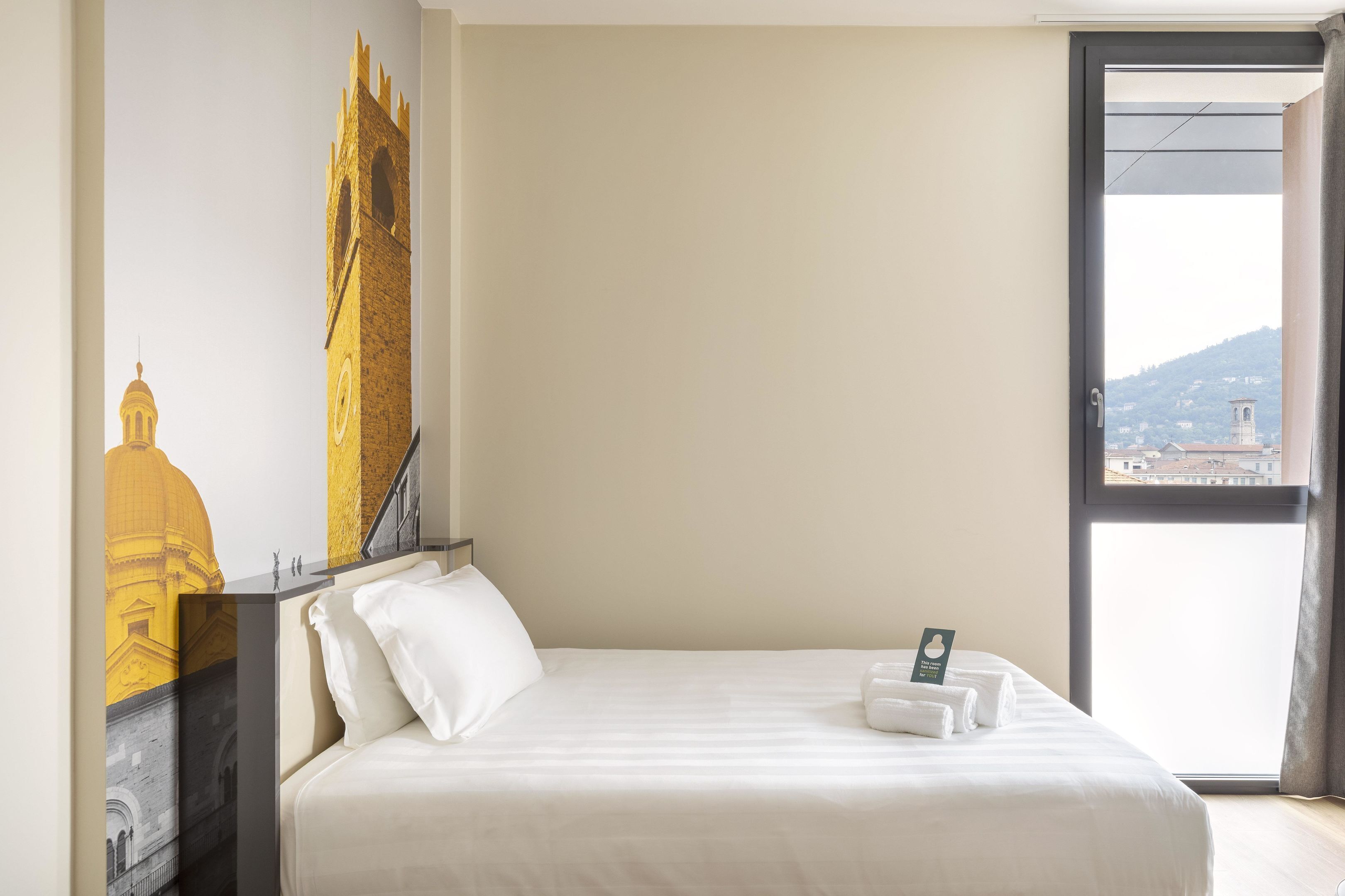 Ett hotellrum med en stor säng, utsikt över en stadsbild genom ett fönster och en väggmålning som föreställer en byggnad.