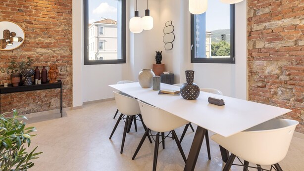 A modern dining area with a white table, black chairs, and a brick wall. There are hanging lights, a vase, and a small sculpture on the table.