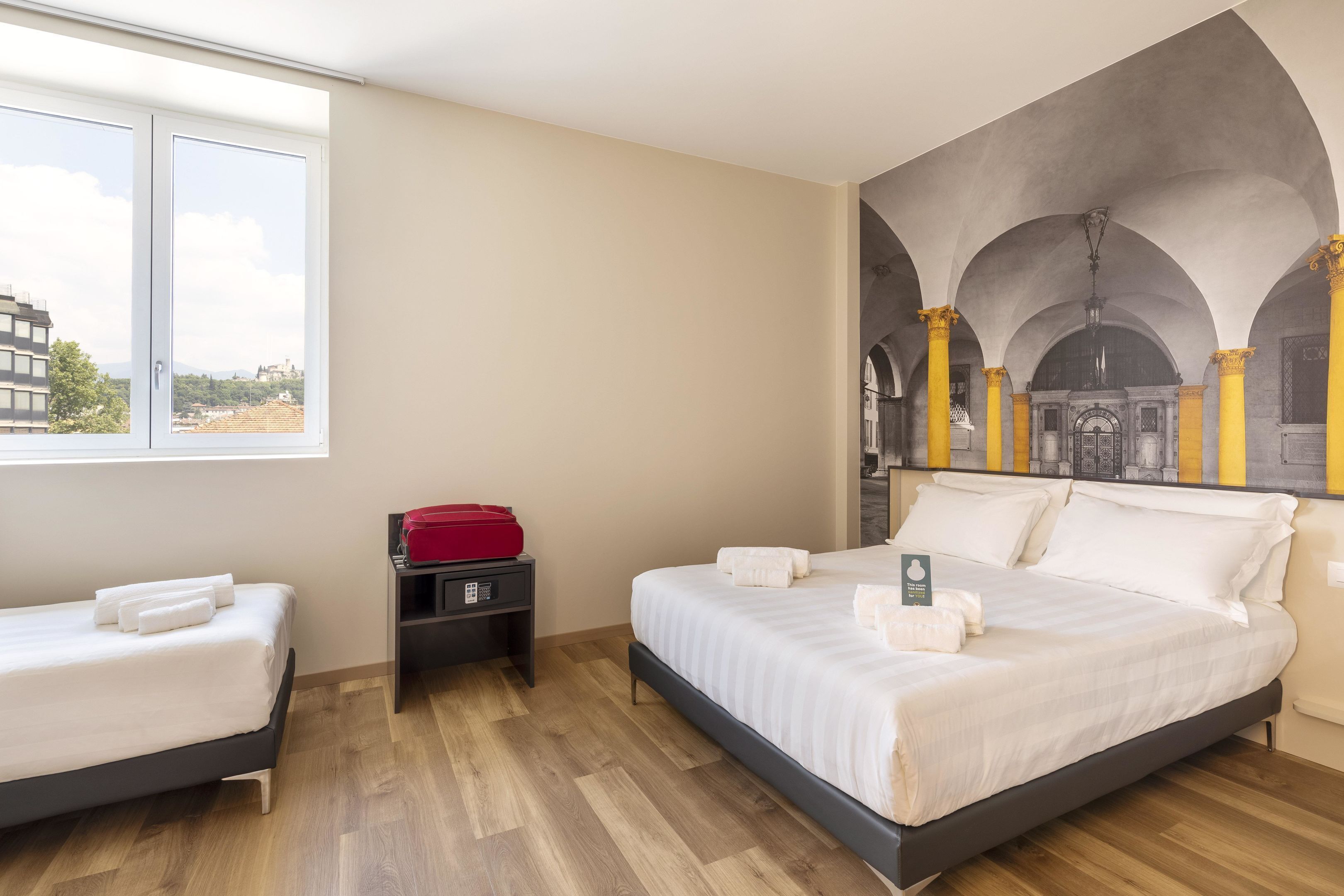 Une chambre d’hôtel avec deux lits, une fenêtre donnant sur la ville et une fresque représentant des arches et des colonnes.