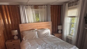 Une chambre à coucher avec une tête de lit en bois, un grand lit avec une literie blanche, deux tables de chevet avec des lampes et une fenêtre avec des rideaux.