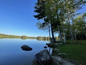 Cena tranquila à beira de um lago, com águas calmas, céu claro e área arborizada com árvores e pedras.