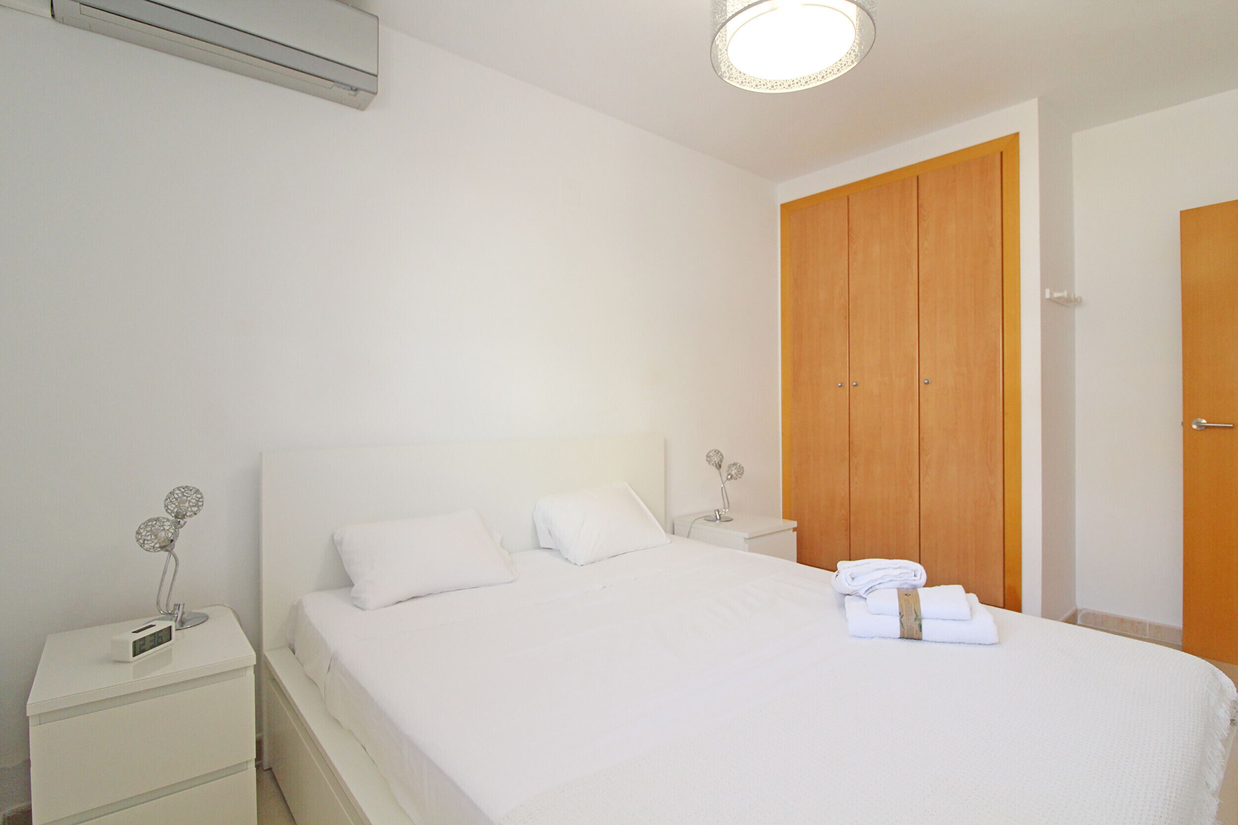 Un dormitorio limpio y minimalista con una cama grande, ropa de cama blanca, un armario de madera y una mesita de noche con lámpara.