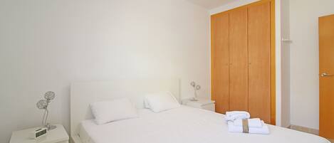 Un dormitorio limpio y minimalista con una cama grande, ropa de cama blanca, un armario de madera y una mesita de noche con lámpara.