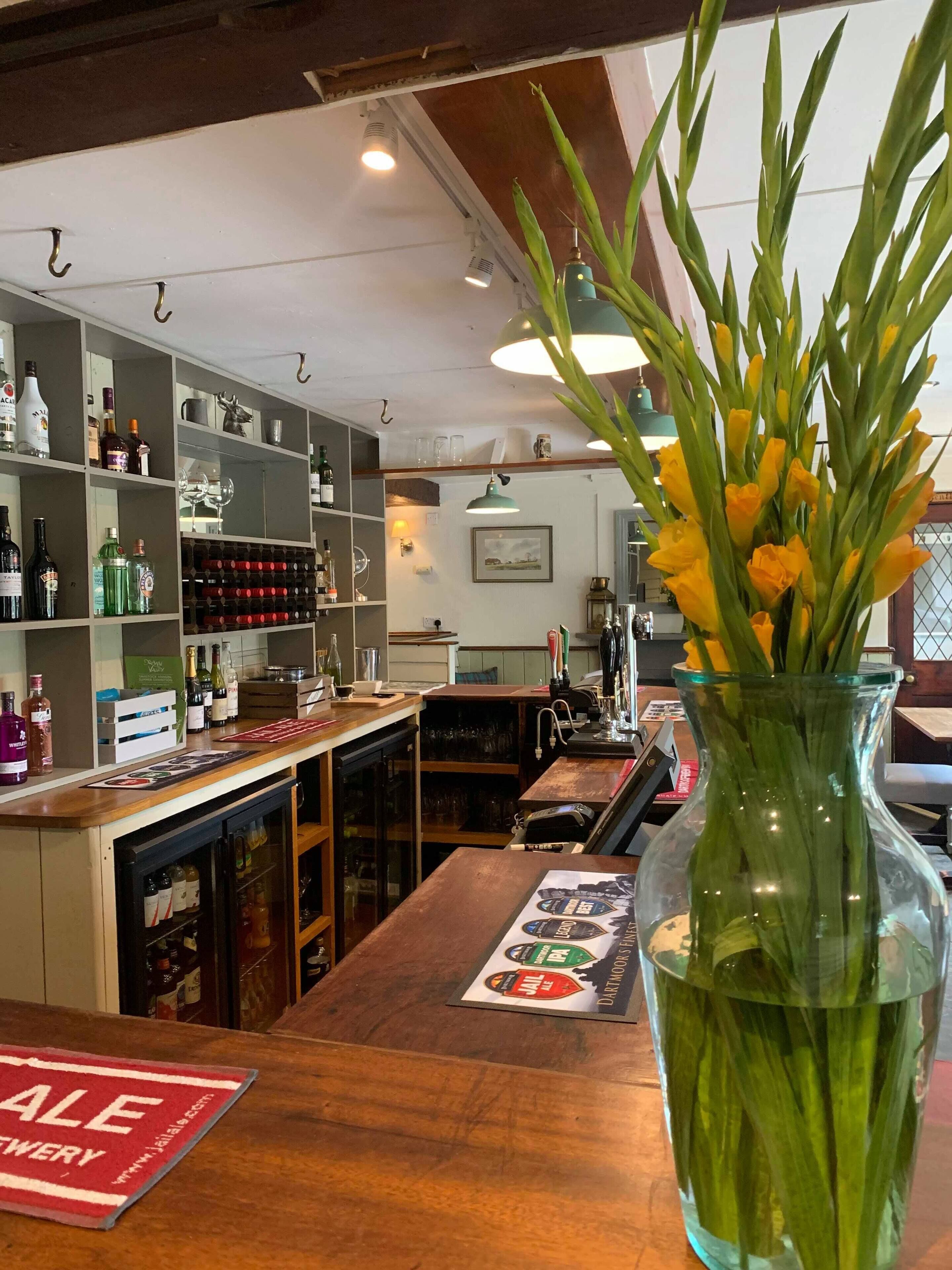 Un espace bar avec des étagères remplies de bouteilles d’alcool, un vase en verre avec des fleurs et une affiche « SOLDES » sur le comptoir.