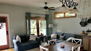 Ein gemütliches Wohnzimmer mit einem dunkelblauen Sofa, einem Holzespeisetisch mit weißen Stühlen und einer Küchenzeile, in der Töpfe hängen.