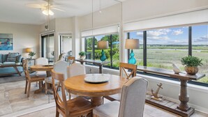 A spacious dining area with a large window offering a view of a field and water. There are several wooden chairs and a round table with a decorative plate.