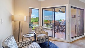 A spacious living area with a sofa, a chair, a small table, and a floor lamp. There is a sliding glass door leading to an outdoor patio with a table and chairs.