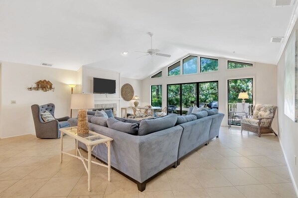 Relax in this spacious living area with a high ceiling and large windows offering a view of the surrounding greenery. The room features a comfortable sofa, a side table, a lamp, a television, and a decorative wall piece.