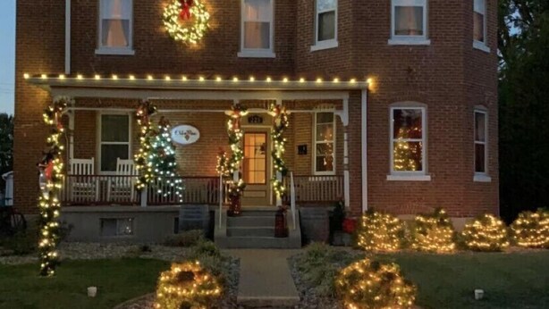 Une maison à deux étages, construite en briques, avec un porche décoré de guirlandes lumineuses et de couronnes de Noël.