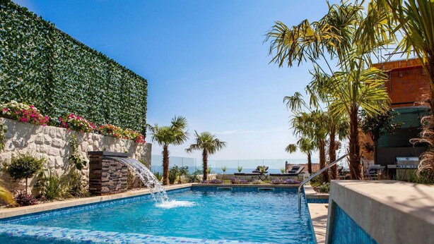 Une piscine agrémentée d’une cascade, entourée de palmiers et bordée d’un mur de pierre orné de fleurs.