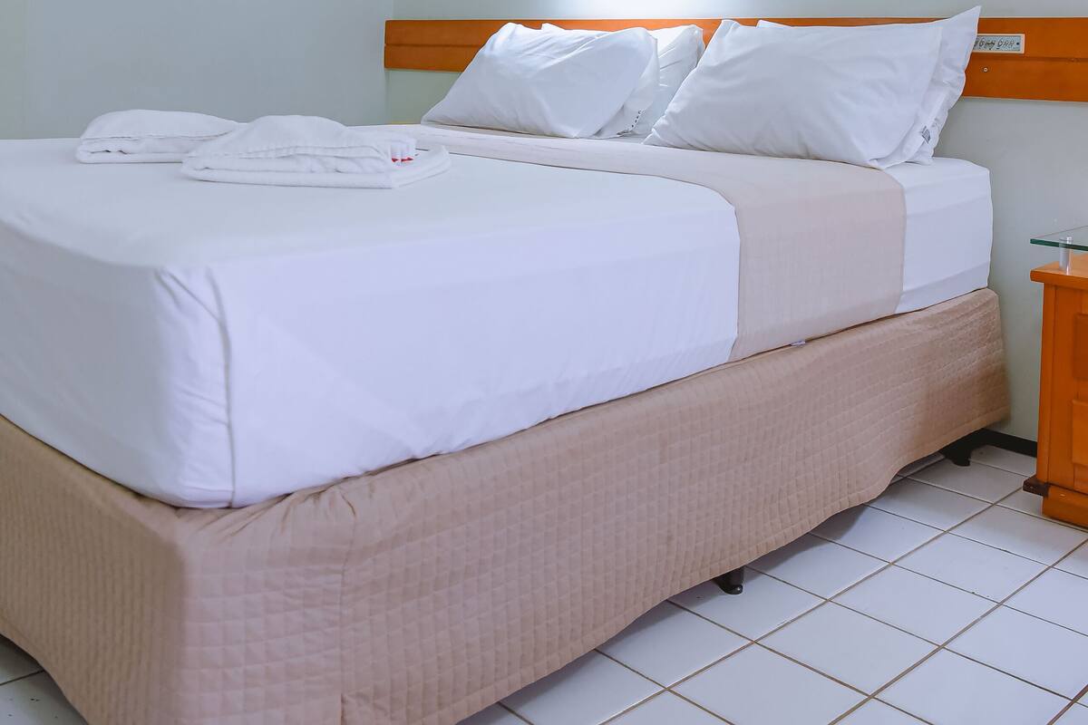 A neatly made bed with white linens and a beige quilted bed skirt, a wooden headboard, a bedside table with a lamp, and a wall-mounted light fixture.