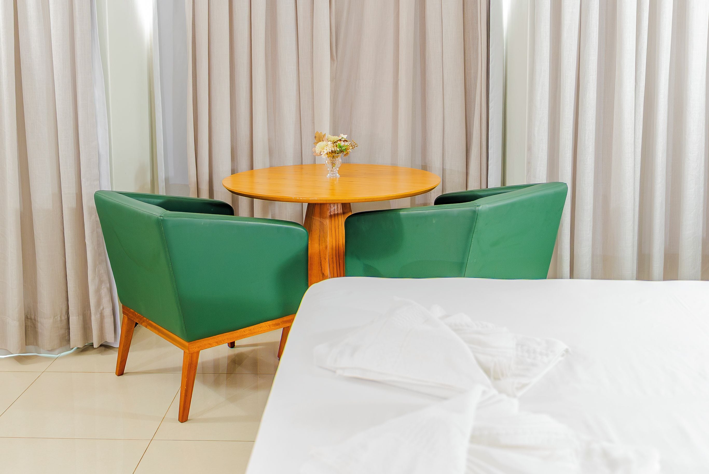 A hotel room with a bed, a round wooden table with two green chairs, and a vase with flowers.