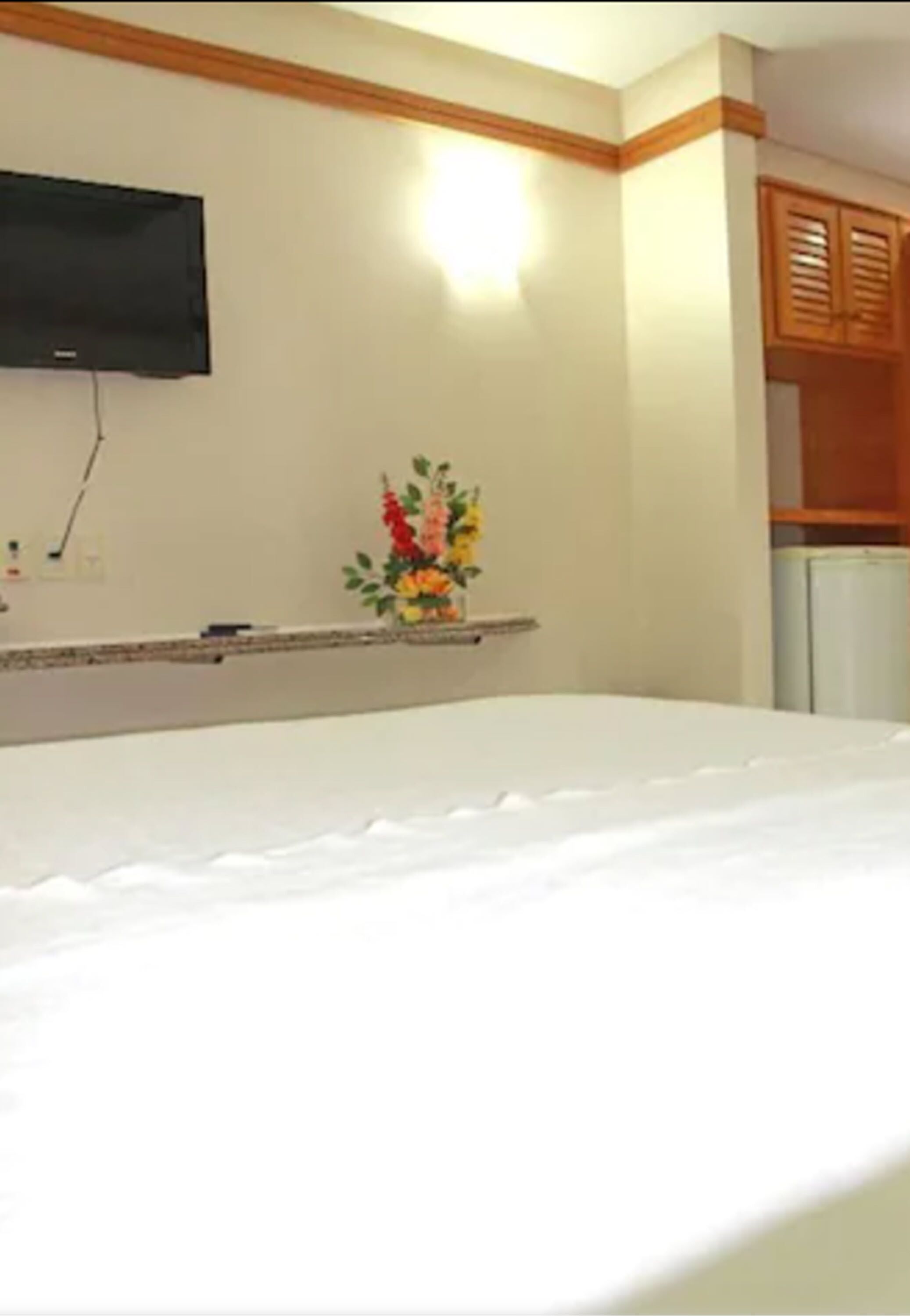 Una habitación de hotel con cama, un televisor de pantalla plana montado en la pared, una mesita con un jarrón de flores y armarios de madera.