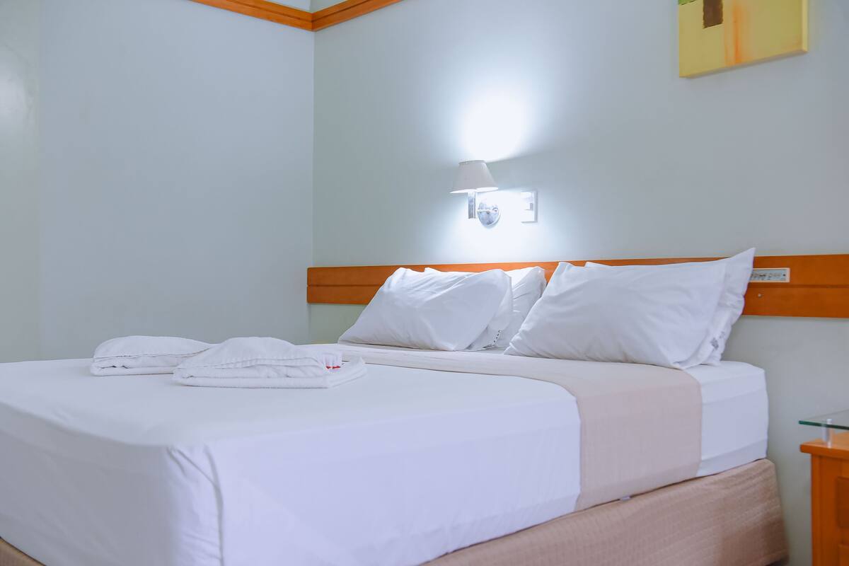 A neatly made bed with white linens and pillows, a bedside table with a lamp, and a wall-mounted light fixture.