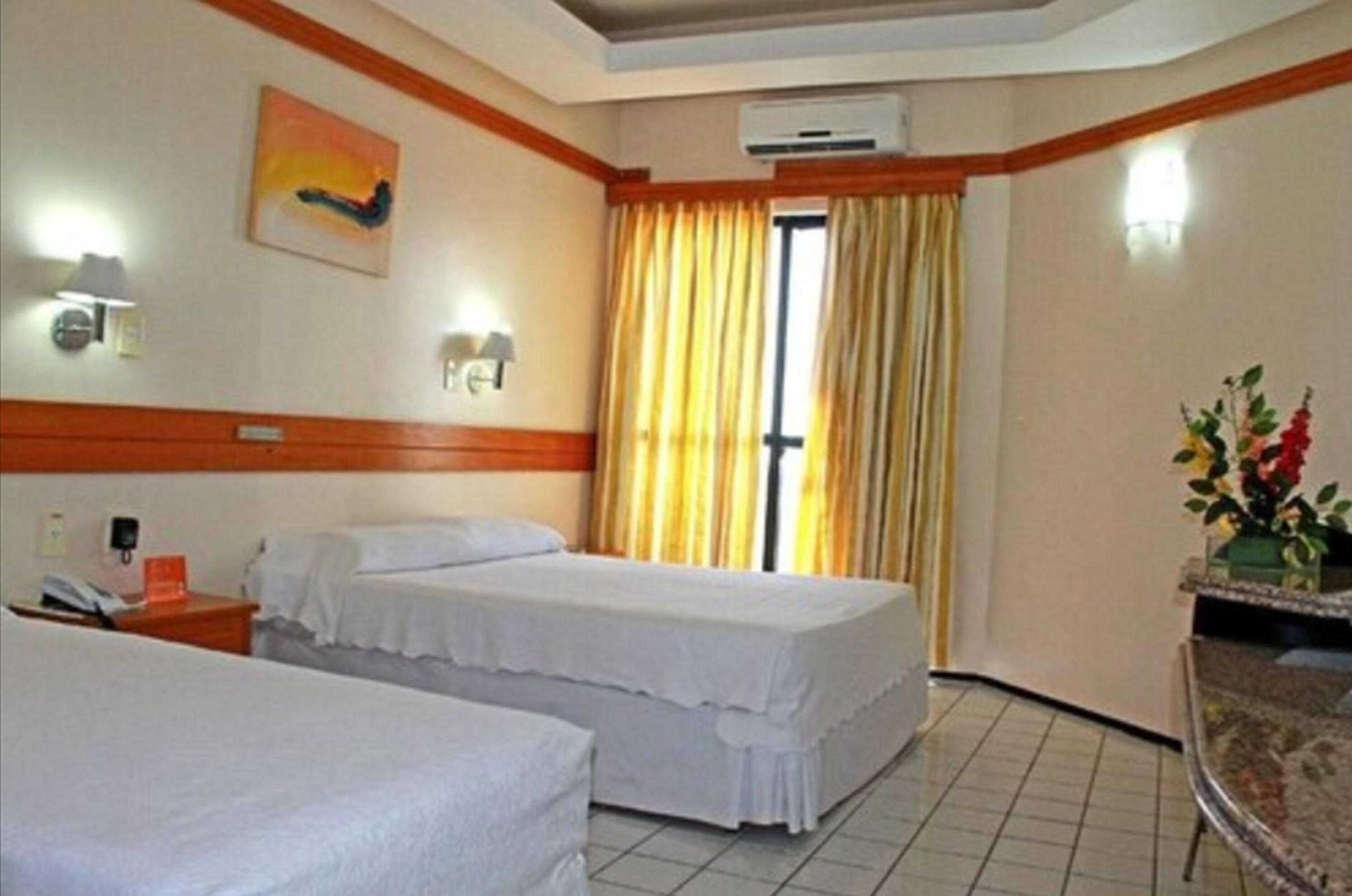 A hotel room with two beds, a desk, a chair, a vase with flowers, and a painting on the wall.