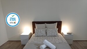 A neatly made bed with a grey and white patterned bedspread, two matching pillows, and a rolled white towel on the bed. There are two bedside tables with lamps, one on each side of the bed. The room has wooden flooring and white walls. - Alojamento 2 Dos Santos (Pinhão)