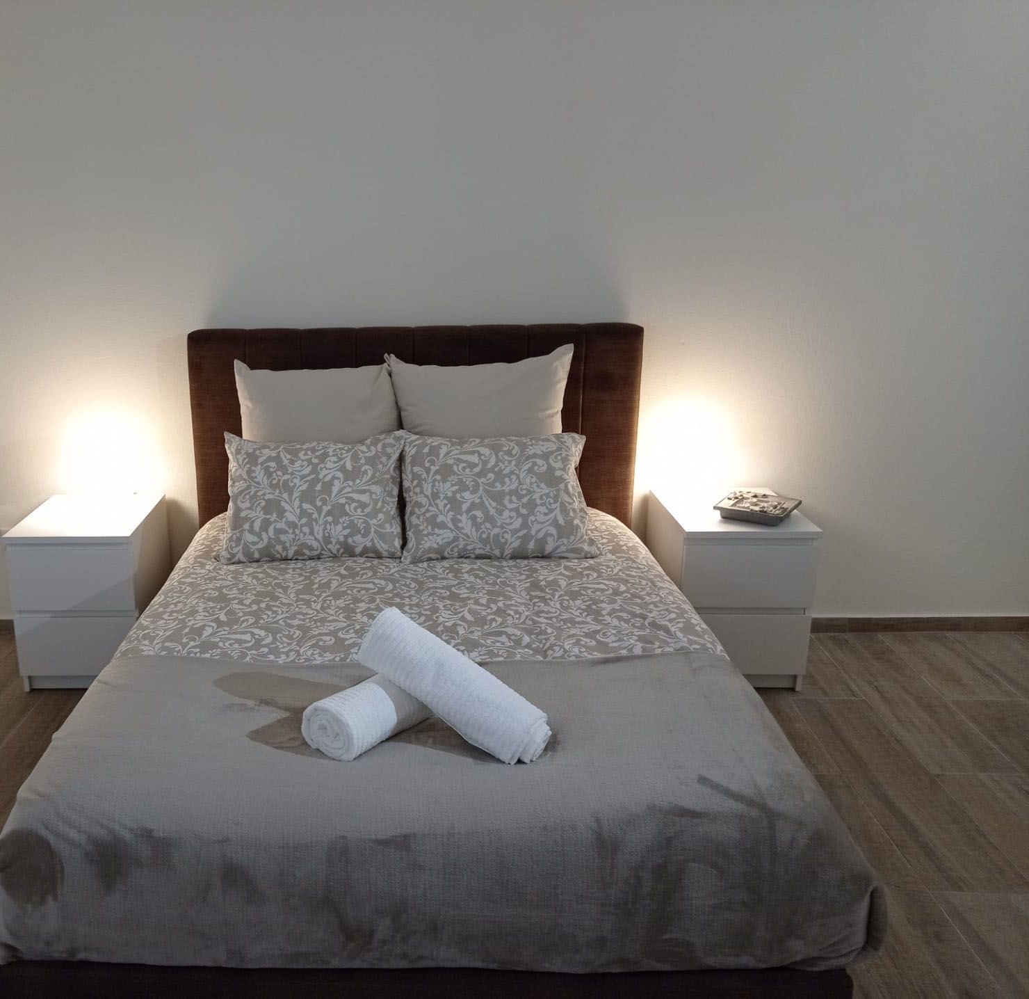 Un lit bien fait, recouvert d’une couvre-lit à motifs gris et blancs, avec deux oreillers assortis et une serviette blanche roulée posée sur le lit. Deux tables de chevet avec des lampes se trouvent de chaque côté du lit. La pièce est dotée d’un plancher en bois et de murs blancs.