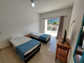 Quarto com duas camas, uma escrivaninha, uma lâmpada no teto e uma janela com vista para edifícios e vegetação.