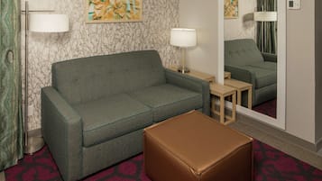 A hotel room with a grey sofa, a brown ottoman, two bedside lamps, a mirror, and a patterned wall.