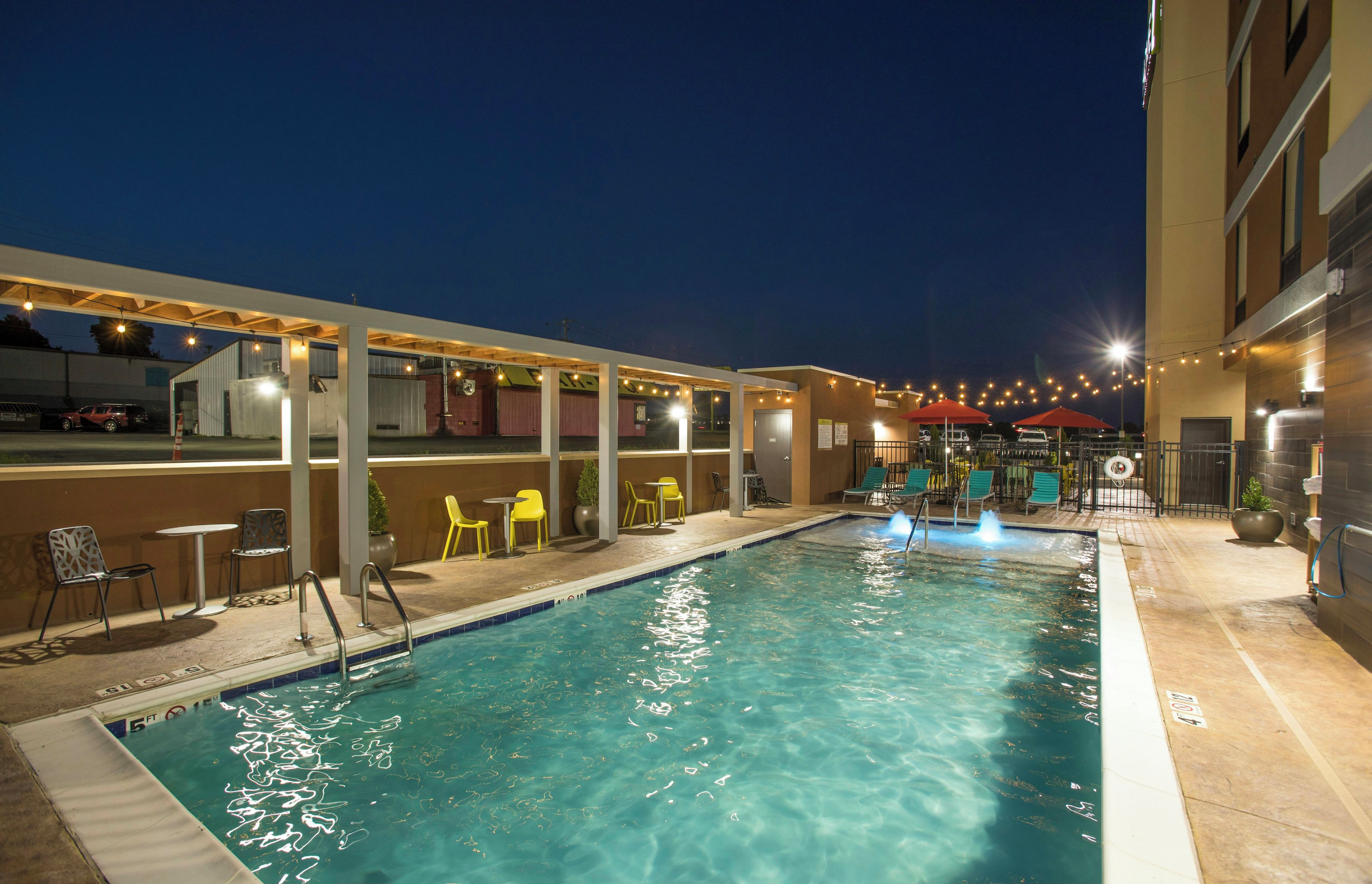An indoor swimming pool with a clear water surface, surrounded by yellow and green chairs, and a covered seating area with string lights.