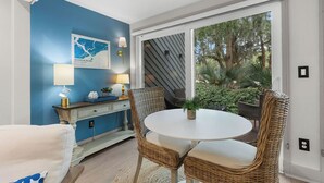 A room with a blue accent wall, a white round table, wicker chairs, a small cabinet with a lamp, and a view of trees through a sliding glass door. - 1 BR/1.5 BA Villa! Amenity access included. NO pets (Seabrook Island)