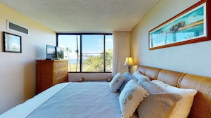 A hotel room with a large bed, a television on a wooden dresser, a window with a view of palm trees and the ocean, and a painting on the wall. - Amazing Oceanview Beachfront Getaway Close to Everything! (Kihei)