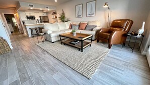 Uma sala de estar moderna com um sofá, mesa de centro e uma poltrona de couro marrom. É possível ver uma cozinha ao fundo, com micro-ondas e frigorífico.