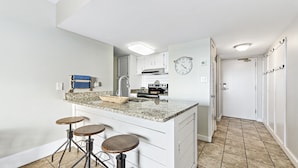 Une cuisine moderne dotée d’un comptoir en granit, d’appareils électroménagers en acier inoxydable et d’un four encastré. On y trouve trois tabourets près de l’îlot central et une horloge au mur.