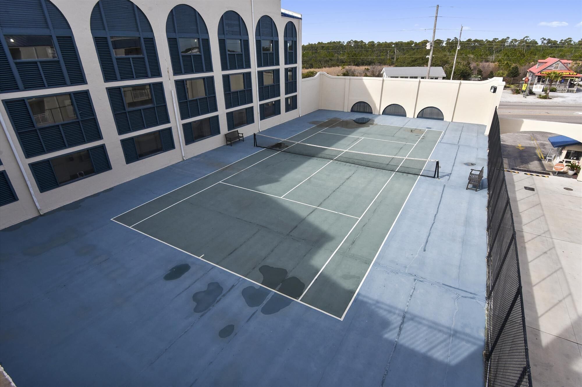 An outdoor tennis court with a net, surrounded by a building with multiple windows and a fence.