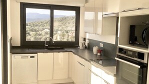 A modern kitchen with white cabinets, a built-in dishwasher, a double oven, and a window offering a view of a hilly landscape.