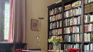 A library with a wooden table, two chairs, a bookshelf filled with books, a vase with flowers, and a red bowl on the table.