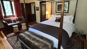 A four-poster bed with a dark wooden frame, a bench at the foot of the bed, a bedside table with a lamp, and a window with red curtains.