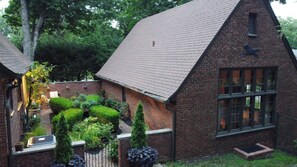 Traditional building with a steeply pitched roof and a large window, set amidst a well-mainters garden with shrubs and a small tree.