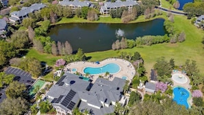 An aerial view of a residential area with a large swimming pool, a tennis court, and a pond.