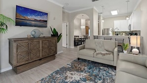 A living room with a sofa, a TV mounted on the wall, a wooden cabinet, and a kitchen area in the background.