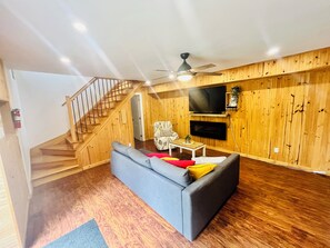 A living room with a grey sofa, wooden flooring, a staircase, a ceiling fan, and a mounted TV.