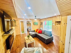 A modern living room with a grey sofa, wooden flooring, a ceiling fan, and a television on a wooden stand.