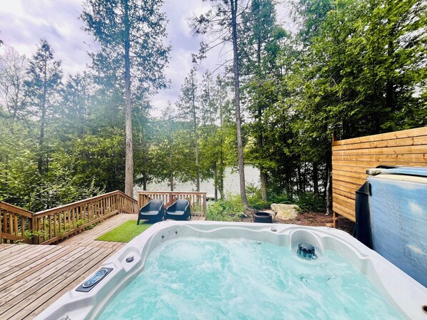 A hot tub surrounded by a wooden deck, with a view of a lake and trees.